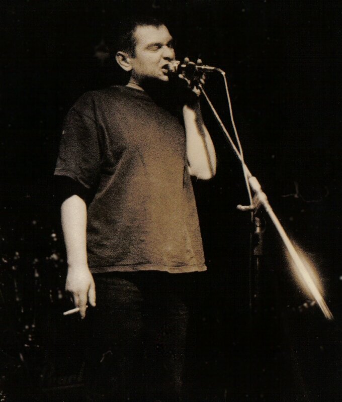 Zdjęcie przedstawia śpiewającego mężczyznę w średnim wieku. Mężczyzna stoi, w lewej ręce trzyma statyw z mikrofonem. W prawej ręce ma palącego się papierosa. Jest ubrany w podkoszulek i dżinsowe spodnie.