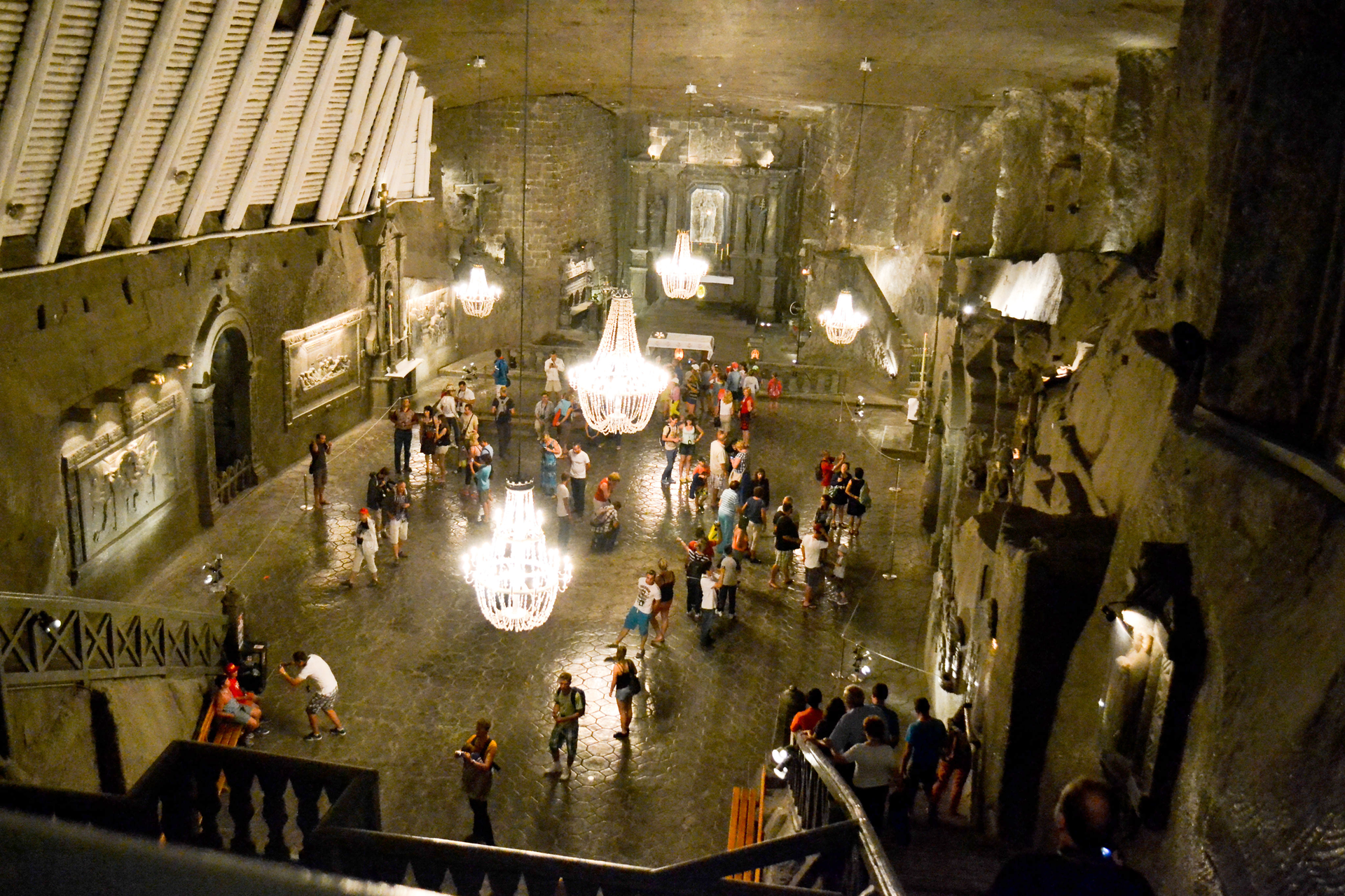 Fotografia wnętrza kaplicy św. Kingi w Wieliczce. Widoczna wysoka sala, oświetlona przez białe kryształowe żyrandole. W kaplicy znajdują się zwiedzający turyści.  Blask kryształowych żyrandoli pada na rzeźbioną posadzkę. Na końcu sali znajduje się ołtarz, a z przodu schody prowadzące do kaplicy.