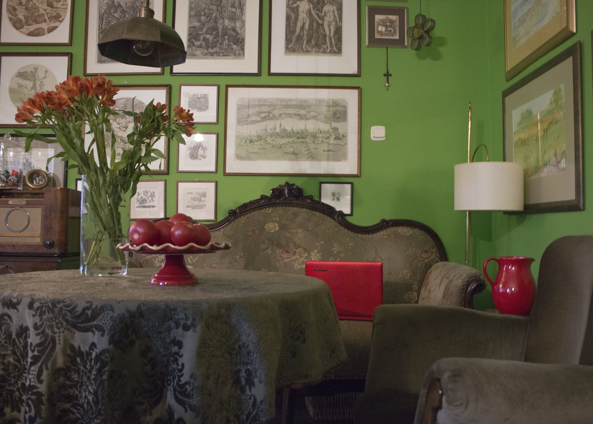 Ilustracja przedstawia zdjęcie wnętrza pokoju urządzonego w starym stylu. Na pierwszym planie znajduje się okryty brązowym obrusem, okrągły stół, na którym stoi czerwona patera z czerwonymi jabłkami oraz bukiet czerwonych kwiatów w szklanym wazonie. U góry kadru, z sufitu zwisa lampa o miedzianym, szerokim kloszu. Obok stołu stoi brązowy fotel. Na zielonych ścianach pokoju wiszą liczne grafiki oprawione w ramki. W głębi wnętrza, pod ścianą umiejscowiona została stara, stylowa kanapa w kolorze brązowym, na której leży czerwony laptop. Obok niej, w kącie stoi lampa z białym kloszem. Po prawej stronie kadru, za fotelem ustawiony został czerwony dzbanek. 