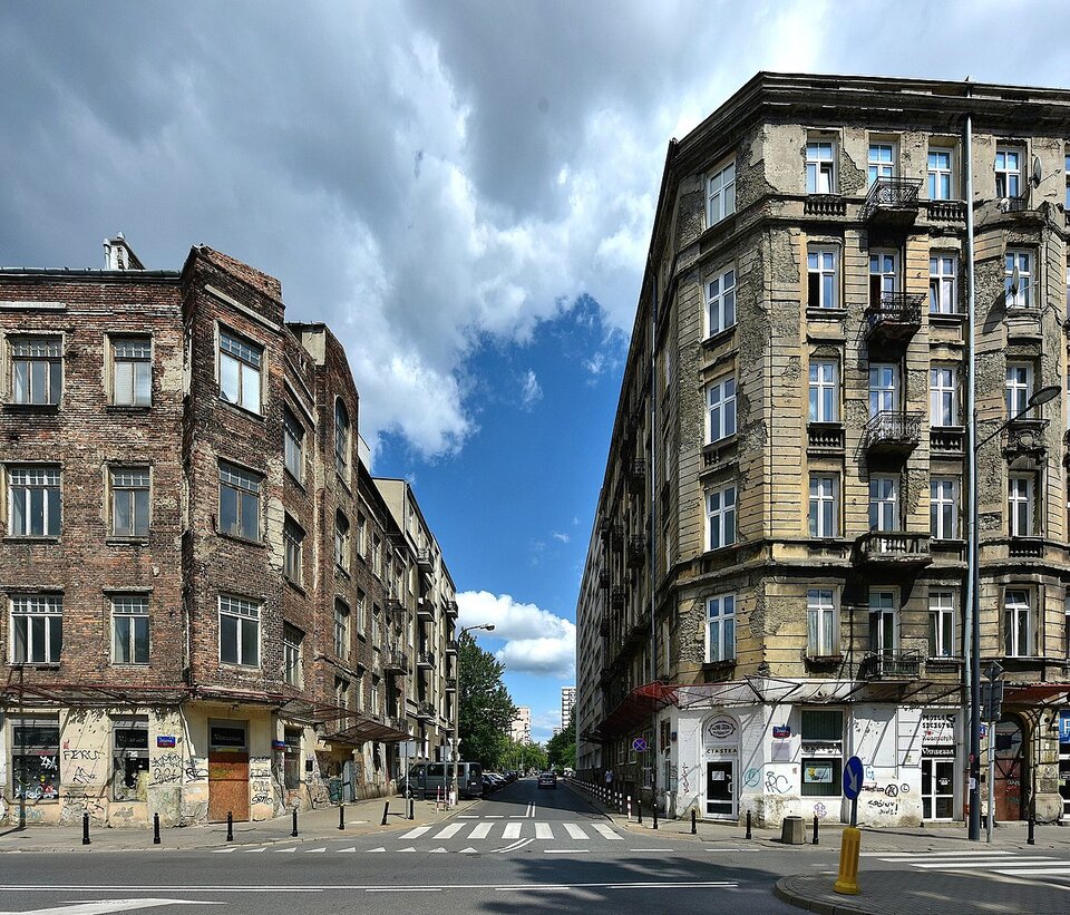 Zdjęcie przedstawia skrzyżowanie ulic w zabudowie miejskiej. Po lewej stronie znajduje się ceglana kamienica z widocznymi śladami graffiti i zniszczonym parterem. Po prawej – tynkowana kamienica z balkonami, również w złym stanie technicznym. Oba budynki mają po kilka kondygnacji. Na ulicy znajduje się przejście dla pieszych, słupki zabezpieczające oraz znaki drogowe. W tle widoczna jest dalsza część ulicy z nieliczną zabudową wielopiętrową. Niebo częściowo zachmurzone.