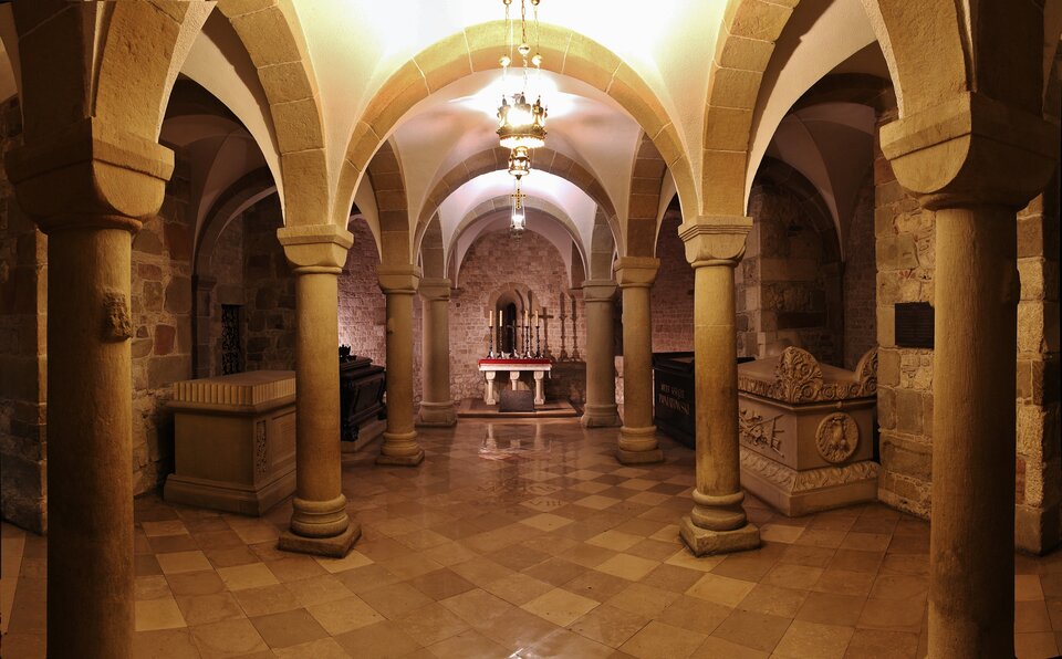 Ilustracja o kształcie poziomego prostokąta przedstawia wnętrze krypty św. Leonarda na krakowskim Wawelu. Na kolumnach, ustawionych w dwóch rzędach, wspiera się sklepienie krzyżowe nakrywające dwie nawy i apsydę krypty. Ściany krypty wykonane są z ciosów kamiennych. Po bokach widoczne są grobowce. W absydzie znajduje się ołtarz.