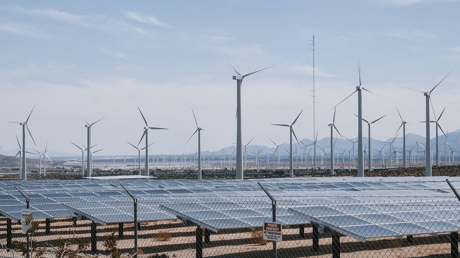 Zdjęcie przedstawia elektrownię wiatrową fotowoltaiczną. Widoczne są potężne wiatraki ustawione jeden za drugim a obok nich ułożono na podwyższeniu panele słoneczne. Teren, na którym znajduje się farma został ogrodzony płotem z siatki stalowej.