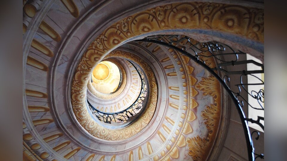 Zdjęcie przedstawia spiralną klatkę schodową. Ukazana jest kuta, metalowa poręcz oraz liczne zdobienia.