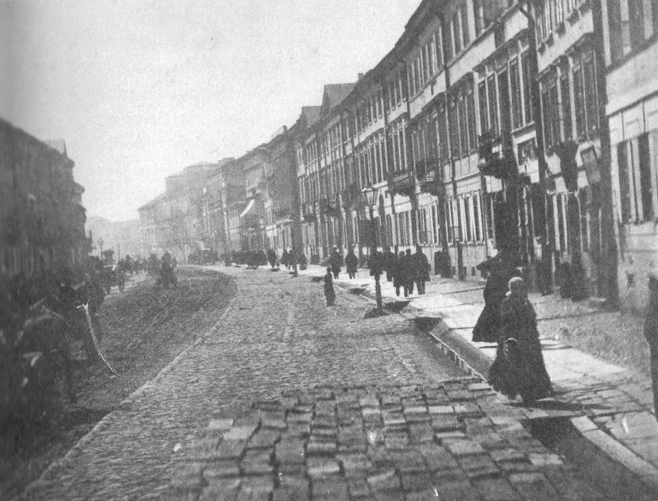 Fotografia przedstawia ulicę skręcającą w lewą stronę. Jej nawierzchnia jest brukowana prostokątnymi, dużymi kamieniami. Wzdłuż traktu stoją kamienice. Na trotuarze ukazani są liczni przechodnie. Po lewej stronie jeżdżą powozy.