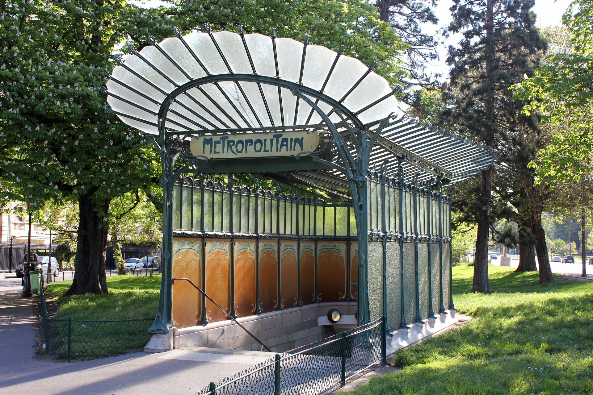 Ilustracja przedstawia fotografię ukazującą wejście do podziemnej stacji metra. Pod zadaszeniem wejścia do stacji wisi tabliczka z napisem „Metropolitan”. Nad wejściem widać szklany dach w kształcie wachlarza. Wejście mieści się w terenie zielonym, otoczone jest drzewami.