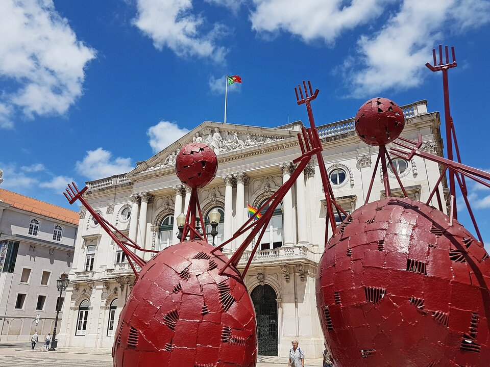 Duża, czerwona metalowa rzeźba przedstawiająca stylizowane postacie z kulistymi głowami i uniesionymi rękami stoi na placu przed okazałym, jasnym budynkiem z kolumnami i zdobioną fasadą. Nad budynkiem powiewa flaga Portugalii, a na balkonie widoczna jest tęczowa flaga. Niebo jest intensywnie niebieskie z białymi chmurami, a na placu spacerują pojedyncze osoby.