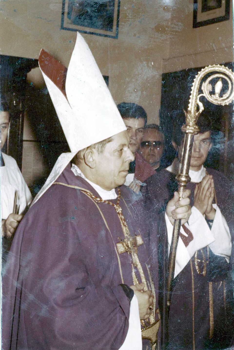 Zdjęcie przedstawia mężczyznę ubranego w szaty liturgiczne. Na głowie ma nakrycie głowy w formie wydłużonej czapki, składającej się z dwóch wysokich, połączonych ze sobą trójkątów, z opadającymi z tyłu dwiema wstążkami, tak zwaną mitrę. W ręku trzyma długą, zdobioną laskę o ślimakowato zwiniętym zakończeniu, tak zwany pastorał. Ubrany jest w sutannę w kolorze fioletowym, na piersiach duży krzyż, wiszący na łańcuchu zawieszonym na szyi.