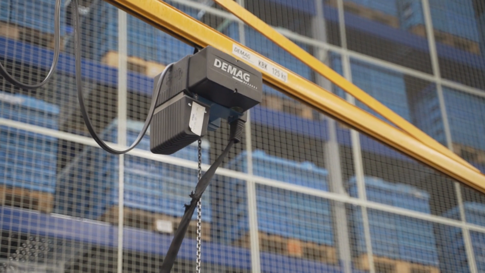 Zdjęcie przedstawia czarną wciągarkę. To urządzenie przenosi się po szynach, które są zawieszone na górze między regałami. Ze wciągarki zwisają łańcuch i pasek, do który zaczepia się towar. Następnie wciągarka podnosi ładunek i przewozi go do miejsca przeznaczenia. W tle jest regał z paletami. Na paletach leży niebieski ładunek.