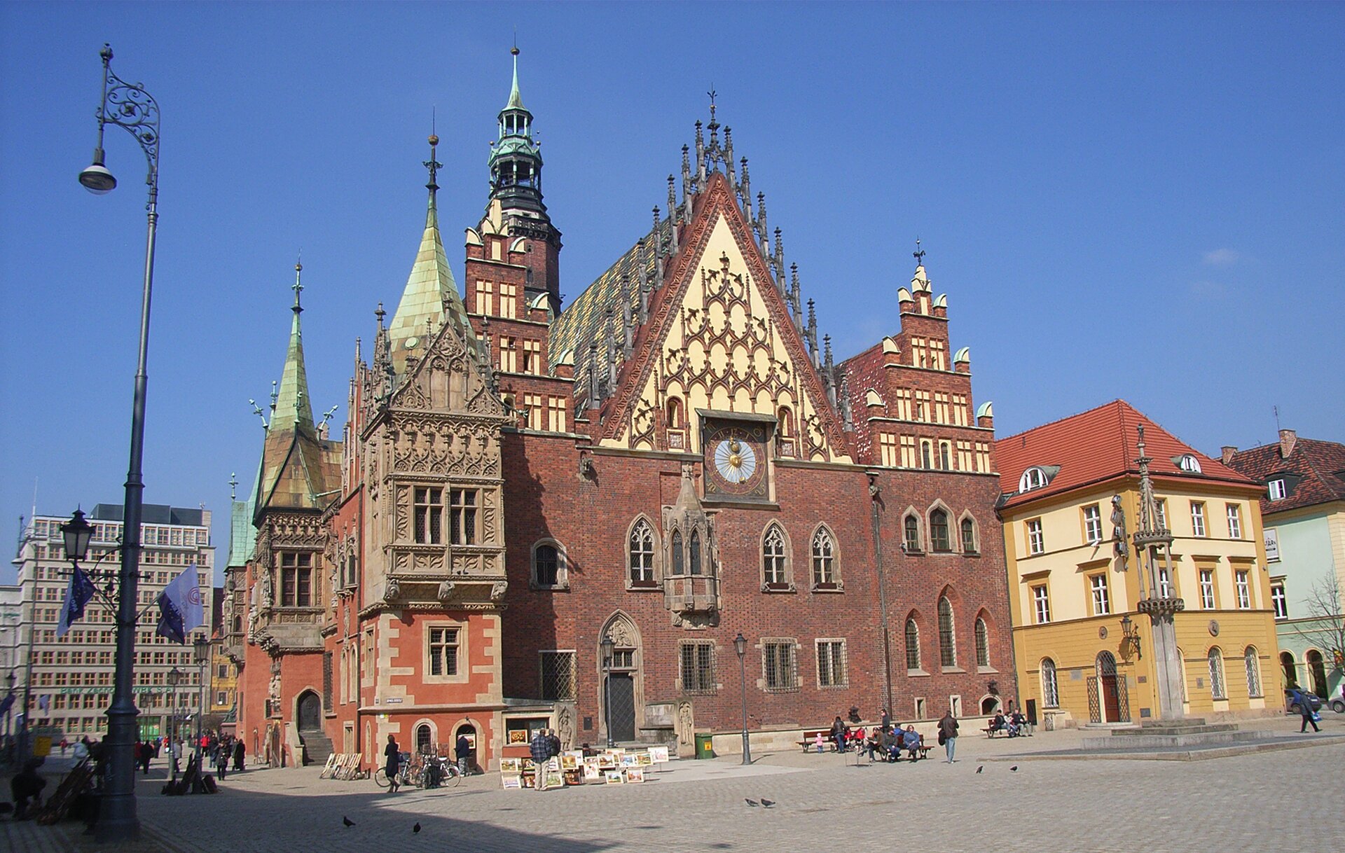 Zdjęcie przedstawia główną elewację gotyckiego ratusza z czerwonej cegły. Jest to dwupiętrowy budynek z wieżą, której fragment widać na drugim planie. Budynek zwieńczony jest trzema szczytami. Środkowy jest otynkowany i bogato zdobiony. Jego dach pokrywają ceramiczne dekoracje, a krawędź zwieńczona jest sterczynami. Boczne są schodkowe z prostokątnymi blendami. Na ścianie frontowej, w centralnej części powyżej drugiego piętra umieszczony jest zegar, a na prawo od niego, na wysokości pierwszego piętra nieduży wykusz. Okna w części środkowej są na pierwszym piętrze prostokątne, na drugim ostrołukowe. Okna  na prawo od głównej części na obydwu piętrach są ostrołukowe. Po lewej stronie znajduje się ostrołukowe wejście do budynku. Po lewej stronie jest przybudówka z wykuszami. Na prawo od ratusza stoją kamienice. Po rynku przed ratuszem spacerują ludzie.
