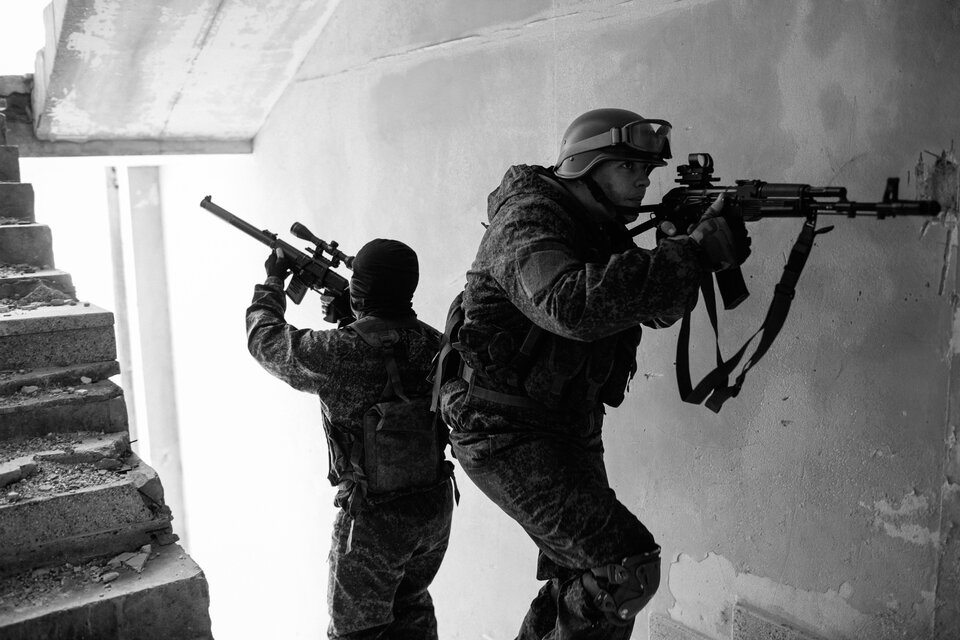 Czarno-biała fotografia przedstawiająca dwóch uzbrojonych żołnierzy lub członków jednostki specjalnej prowadzących operację w zrujnowanym budynku. Obaj ubrani są w taktyczne mundury maskujące i wyposażeni w kamizelki taktyczne. Jeden z nich, noszący hełm, celuje karabinem szturmowym w stronę ściany, podczas gdy drugi, w kominiarce, zabezpiecza schody, trzymając karabin z lunetą.