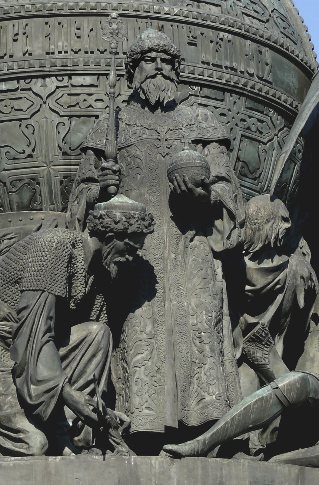 Kliknij, aby powiększyć Zdjęcie przedstawia zbliżenie na fragment szarego pomnika. W centralnej części znajduje się stojący mężczyzna w średnim wieku z dłuższą brodą i wąsami. Na głowie ma czapkę zakończoną kożuchem i ubrany jest w długą, zdobioną szatę. W dłoniach trzyma berło oraz jabłko królewskie. Po obu jego stronach klęczą mężczyźni. Za postaciami widoczny jest fragment kuli.