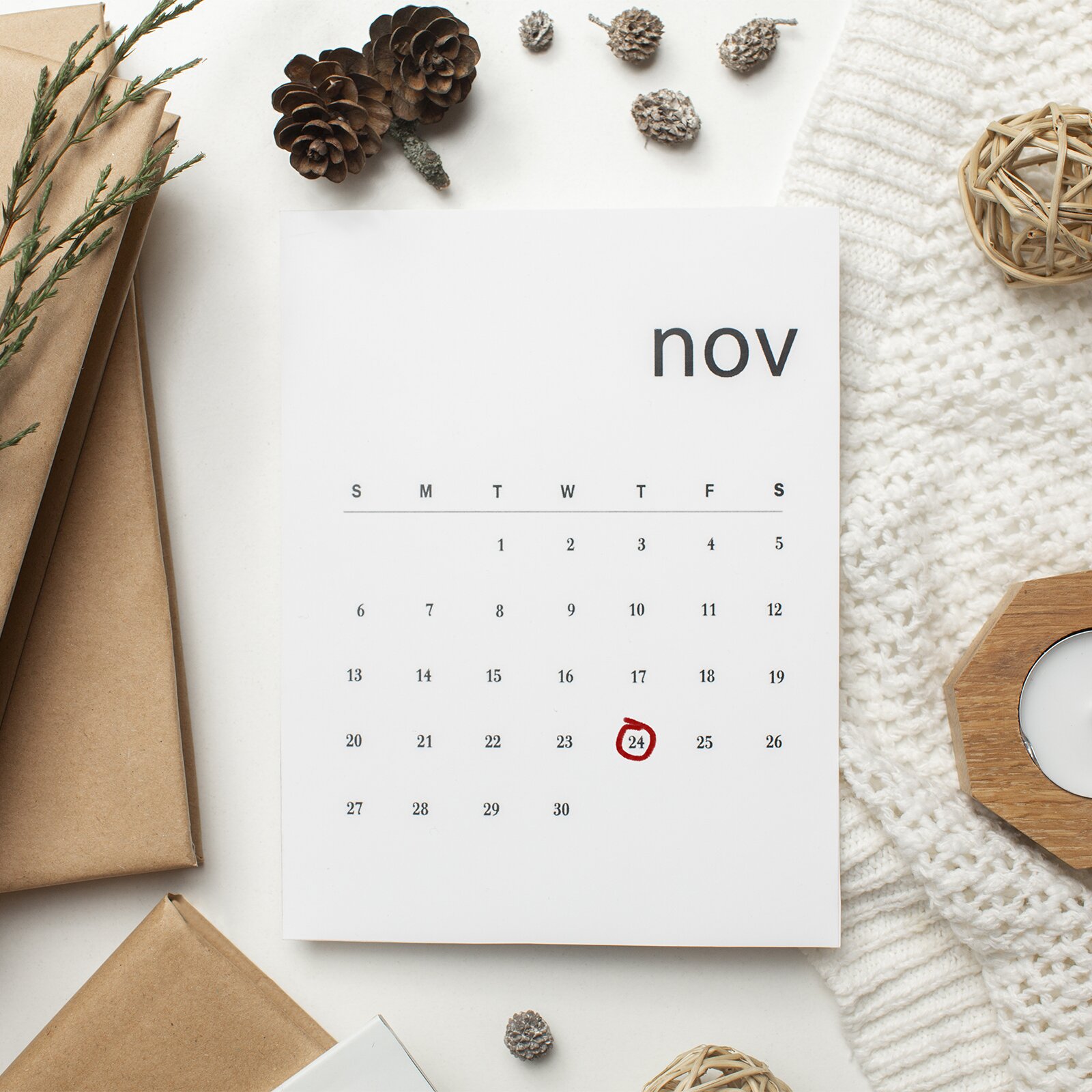 Zdjęcie przedstawia kalendarz z kartkę z miesiącem  listopad. Widoczna jest nazwa miesiąca – nov, skrót od słowa November. Dzień 24 został zaznaczony odręcznie czerwonym kółkiem. Wokół kalendarze widoczne są papierowe torebki, gałązka jałowca, duże i małe szyszki, fragment białej dzianiny, ażurowa kula ozdobna wykona gałązki.