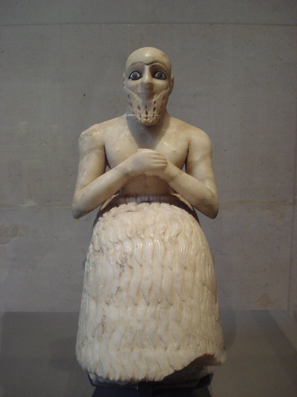 Zdjęcie posągu przedstawiającego siedzącego na tronie masywnego mężczyznę z szeroko otwartymi oczyma i długą dekoracyjnie wyrzeźbioną brodą. Skierowany jest przed siebie. Ręce ma ugięte w łokciach i wsparte na kolanach, a dłonie trzyma skrzyżowane na piersi. Ubrany długą tunikę zasłaniającą nogi. Spod niej widoczne są wystające palce. Na przedniej stronie szaty i na tylnej części tronu widoczne są inskrypcje. 