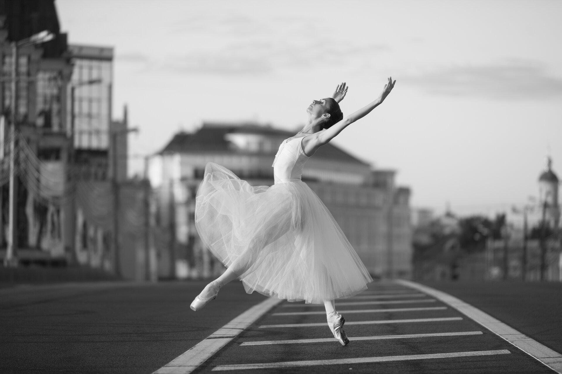 Ilustracja przedstawia baletnicę. Ubrana jest w typowy strój baletowy, czyli bluzkę na ramiączkach, tiulową spódnicę oraz baletki. Kobieta tańczy na ulicy, uchwycony został moment podskoku baletnicy. W tle widoczne są budynki. Fotografia jest czarno-biała.