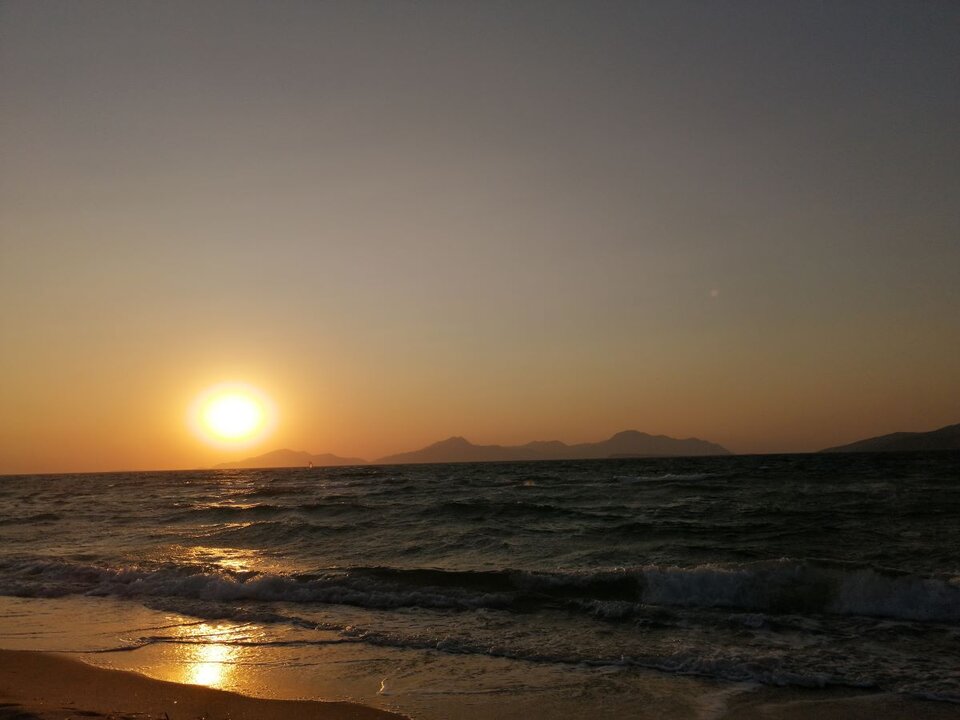 Ilustracja przedstawia fotografię zachodu słońca nad morzem. W kadrze o układzie horyzontalnym znajdują się: do połowy wysokości kadru - morze, powyżej niebo, z lewej strony tuż nad taflą wody, na horyzoncie widać okrągłe słońce. Światło zachodzącego słońca rozświetla bezchmurne niebo oraz morskie fale. 
