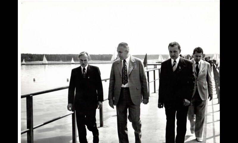 Zdjęcie przedstawia grupę mężczyzn w garniturach, którzy idą przez most. Przy barierkach mostu powiewają flagi. W tle po wodzie płyną żaglówki.