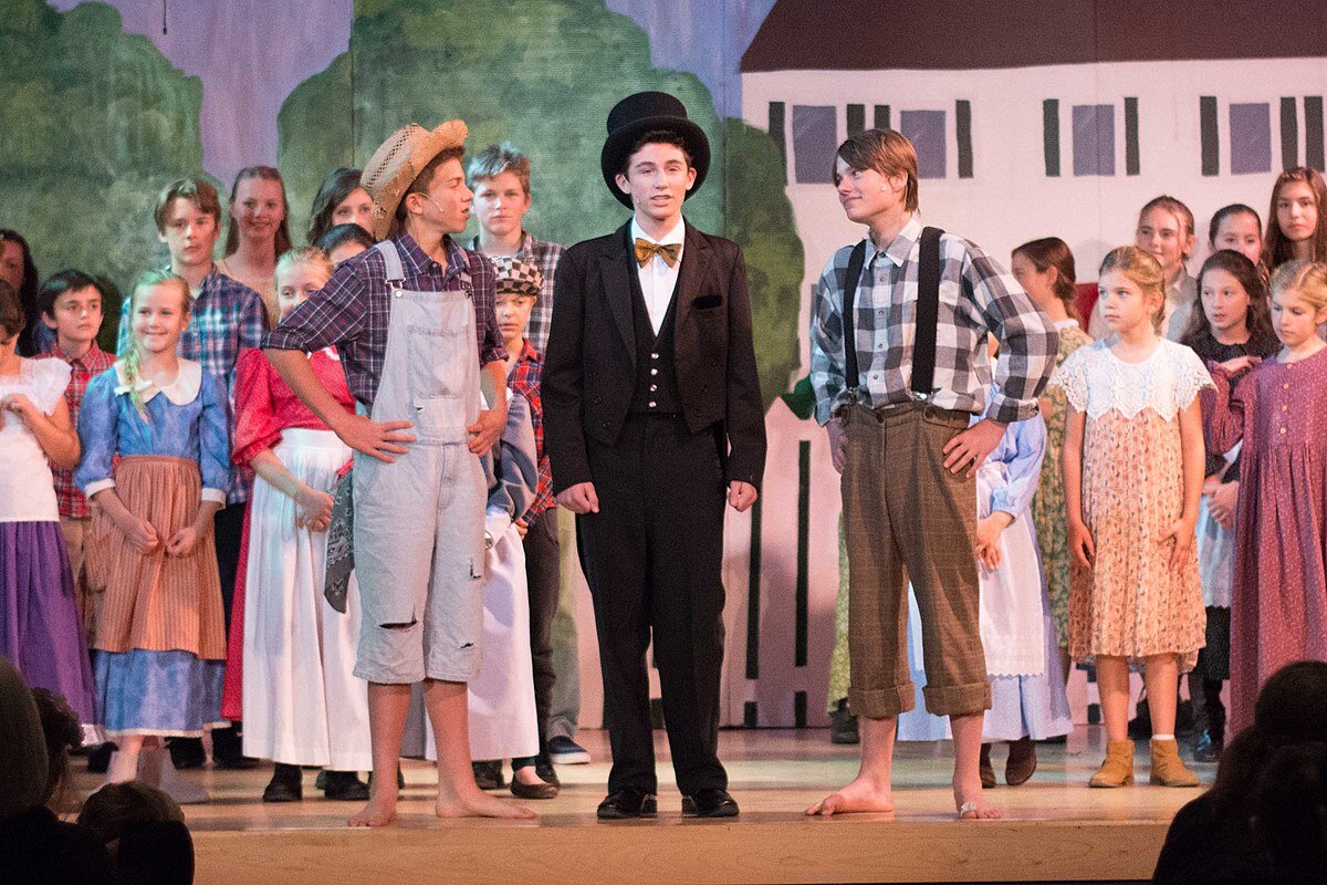 Kliknij, aby powiększyć Fotografia przedstawia szkolne przedstawienie teatralne pt. „Przygody Tomka Sawyera”. Na pierwszym planie znajduje się trzech chłopców. Pośrodku stoi chłopiec w czarnym fraku i cylindrze. Po lewej stronie chłopiec w słomkowym kapeluszu, biało‑fioletowej koszuli w kratę, niebieskich ogrodniczkach z dziurami na nogawkach sięgających kolan. Ma bose stopy i opiera dłonie na biodrach. Po prawej stronie stoi chłopiec w koszuli w biało‑czarną kratę, brązowych spodniach na czarnych szelkach. Ma bose stopy i opiera dłonie na biodrach. Za nimi znajduje się tłum dzieci, a w tle widać dekorację z domem, płotem i drzewami.