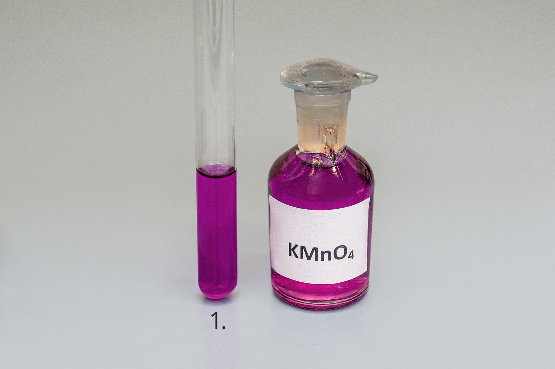 Zdjęcie przedstawia probówkę oraz butelkę ze szklanym korkiem, wypełnione fioletowym roztworem nadmanganianu potasu ka em en o cztery. Roztwór wypełnia 1/3 objętości probówki.