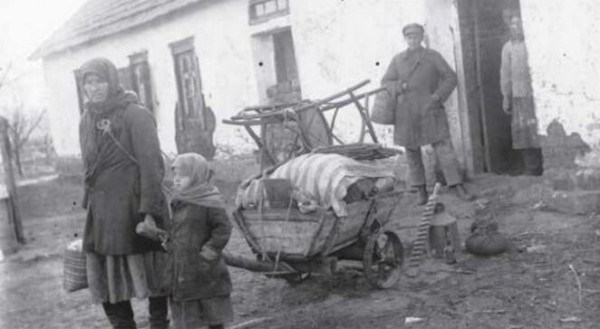 Kliknij, aby powiększyć Na zdjęciu jest kobieta z małą dziewczynką. Ubrane są skromnie, na głowach mają chustki. Kobieta w jednej ręce trzyma wypchaną torbę, w drugiej dyszel od drewnianej taczki. Na taczce leżą rzeczy przykryte materiałem, na tym leży krzesło. Obok kobiety dyszel trzyma dziewczynka. W tle widać budynek. W jego drzwiach stoi osoba. Przed drzwiami stoi mężczyzna.
