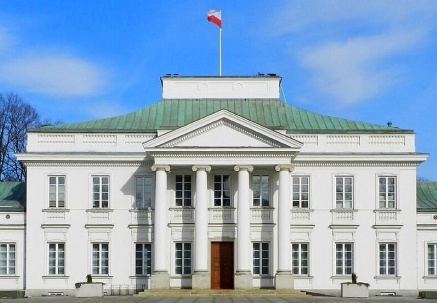 Ilustracja przedstawia „Pałac Belwederski” w Warszawie. Zdjęcie prezentuje budynek od frontu. Jego fasada jest symetryczna. Pośrodku znajduje się zwieńczony trójkątnym tympanonem portyk, wsparty na czterech jońskich kolumnach. Skrzydła boczne to dwie kondygnacje z symetrycznie rozmieszczonymi oknami. Pokryty zielona dachówką dach jest czterospadowy, zwieńczony nadbudówką.