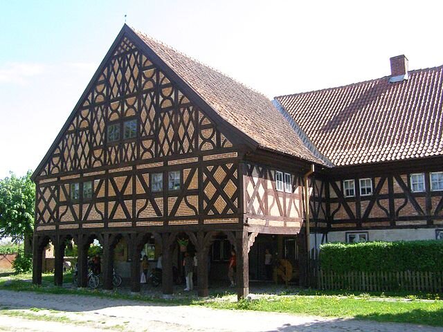 Ilustracja przedstawia drewniany dom. Jego elewacja jest wymalowana w beżowo‑brązowe wzory. Ma drewniane kolumny podtrzymujące sklepienie nad gankiem. Widać ludzi przed wejściem. Wokół rośnie żywopłot. 