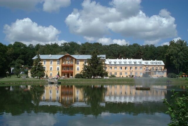 Zdjęcie przedstawia duży budynek w stylu klasycystyczno‑uzdrowiskowym, położony nad brzegiem niewielkiego stawu. Obiekt ma elewację w jasnym, kremowym kolorze, białe okna i dachy pokryte blachą. W centralnej części znajduje się drewniany balkon z zadaszeniem wspartym na słupach. Przed budynkiem widoczna jest fontanna umieszczona na tafli wody, wokół której rozciąga się zadbany trawnik i roślinność ozdobna. W tle rośnie gęsty las. Niebo jest częściowo zachmurzone, z dużymi białymi chmurami. Całość odbija się wyraźnie w wodzie.