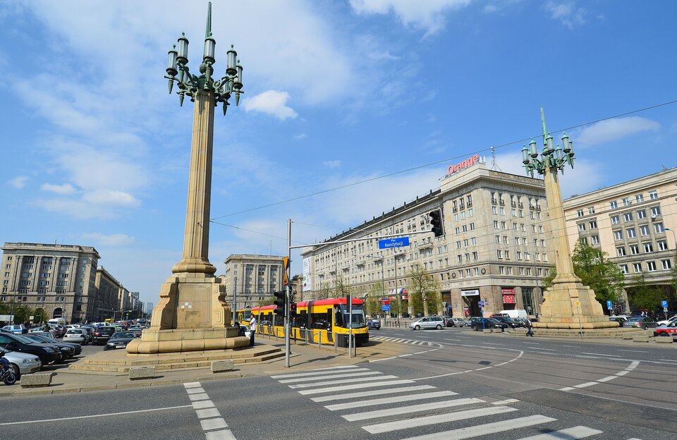 Fotografia przedstawia Plac Konstytucji w Warszawie. Widzimy na nim wysokie ozdobne lampy, wokoło niego przebiega ruch uliczny oraz znajduje się parking, W tle są widoczne budynki w stylu socrealistycznym.