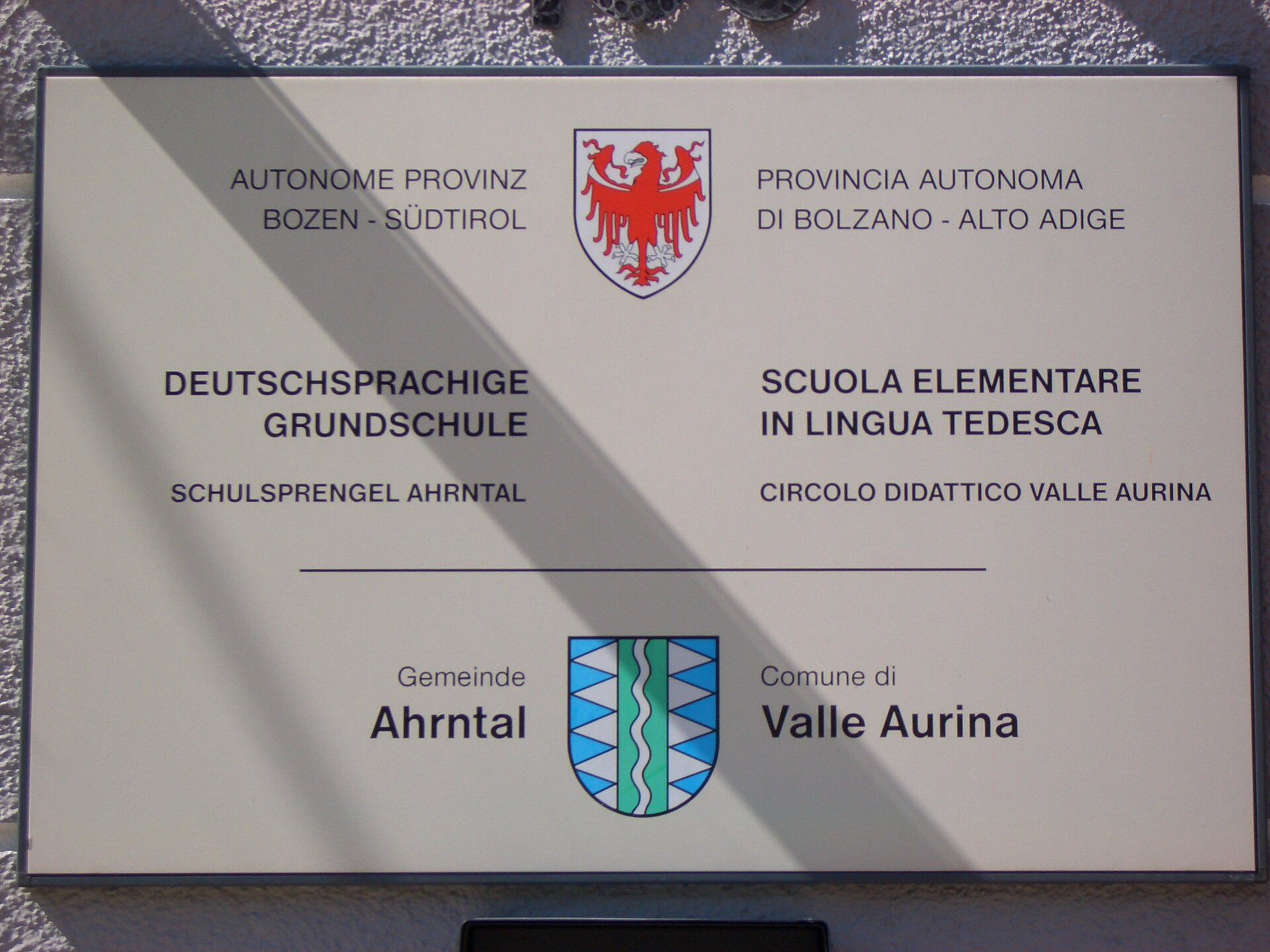 Biała tablica z napisami w języku włoskim i niemieckim. Dwa kolorowe herby, nazwy szkół i nazwy miast.