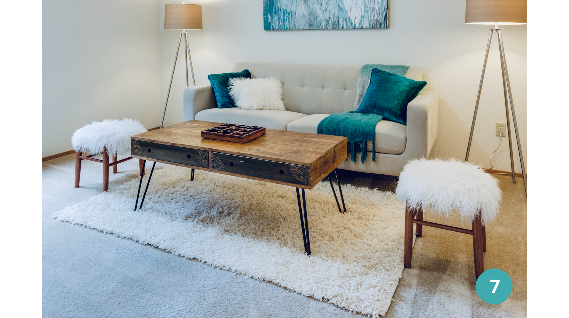 Zdjęcie przedstawia salon. Na środku pod ścianą stoi kanapa, po lewej i po prawej stronie kanapy stoją lampy. Przed kanapą stoi stolik i dwa stołki. Pod stolikiem leży dywan.