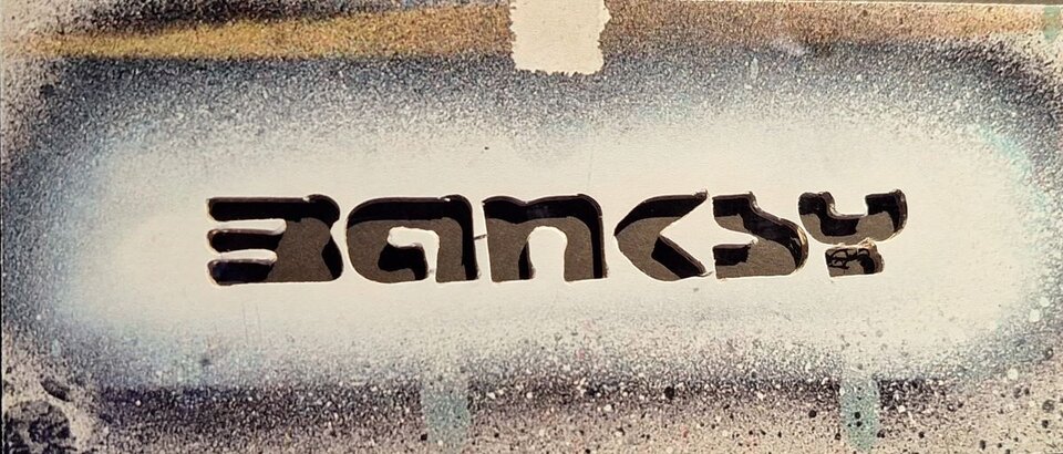 Ilustracja przedstawia szablon napisu "banksy": poziomy karton , na którym są wycięte otwory układające się w napis.