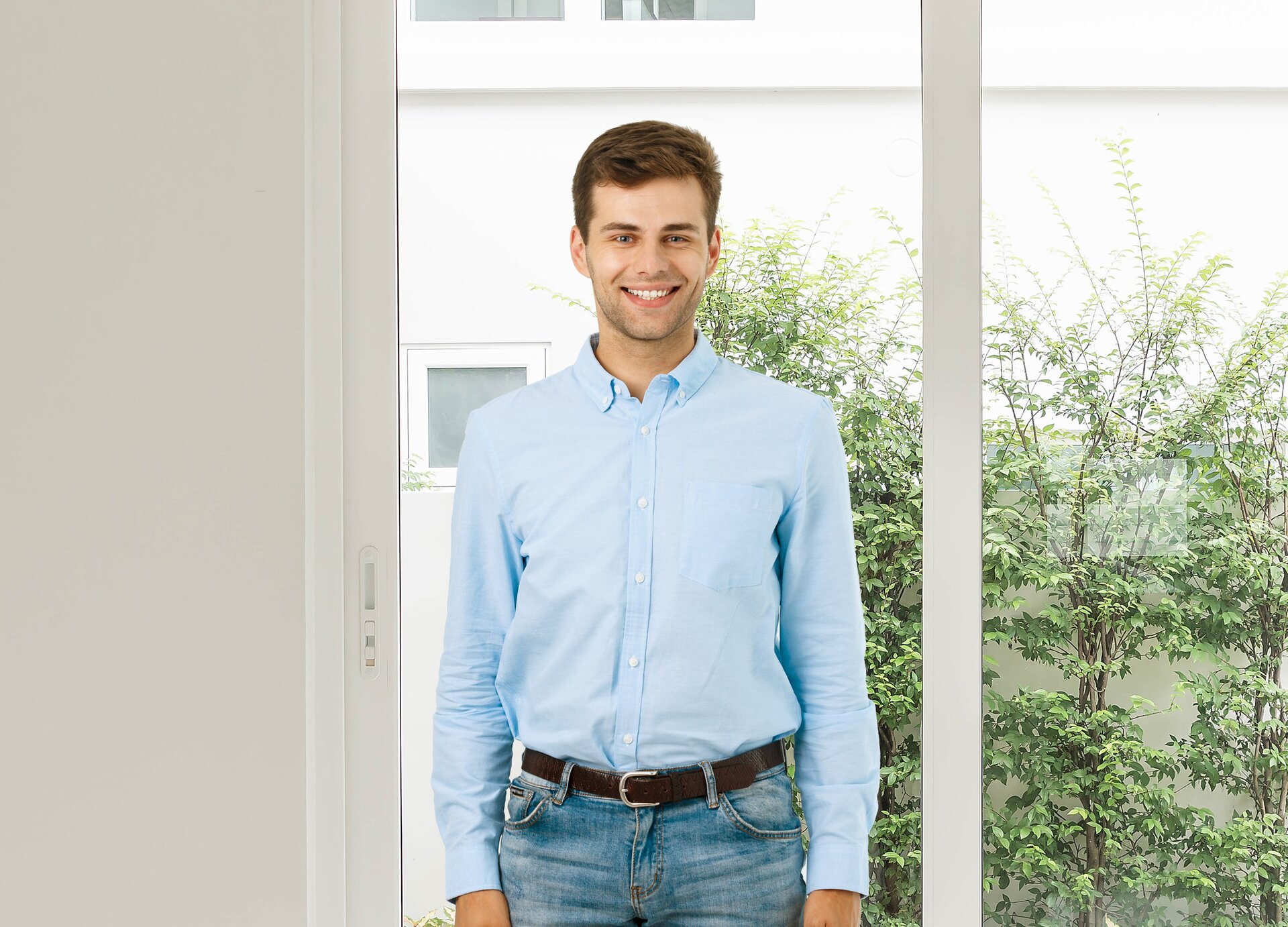 Zdjęcie przedstawia młodego mężczyznę pozującego do zdjęcia na tle okna, za którym widoczny jest zielony krzew. 