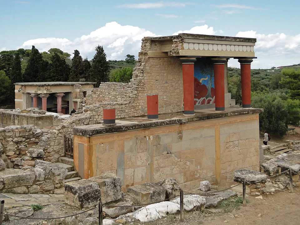 Fotografia przedstawia ruiny pałacu w Knossos na Krecie, dokładnie tzw. północny portyk z kolumnami. Budowla widoczna na pierwszym planie to prostokątny fragment pałacu wzniesiony na kamiennej, jasnobeżowej platformie z dużych, obrobionych bloków. Jej górna część pokryta jest rekonstrukcją portyku wspartego na czterech czerwonych kolumnach rozszerzających się ku górze, z czarnymi głowicami i dolnymi częściami oraz jasnej bazie. Kolumny wspierają płaski dach z ozdobnym fryzem złożonym z rytmicznie ułożonych białych, kołowych dekoracji na ciemnym tle. Ściany budowli wykonane są z jasnego kamienia. Na pierwszym planie, poniżej budowli, znajdują się nieregularne, kamienne pozostałości partii pałacu - bloki i fragmenty murów. W tle po lewej stronie znajduje się kolejna część pałacu z trzema czerwonymi kolumnami, również zrekonstruowanymi, oraz ścianami o podobnej kolorystyce – jasnobeżowej z ciemniejszymi elementami. Budowla otoczona jest roślinnością. Całość ukazana jest na tle błękitnego nieba.