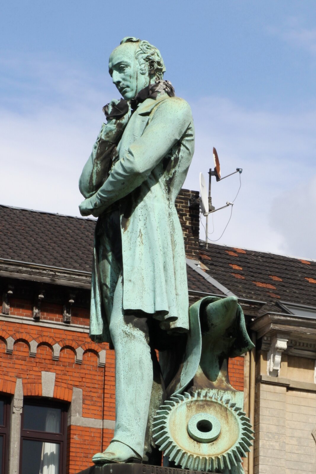 Na zdjęciu pomnik mężczyzny,  trzymającego prawą rękę pod brodą. Włosy zaczesane do tyłu, ubrany w płaszcz do kolan. Ma długie , dość wąskie spodnie opierające się na butach. Lewą nogę ma wysuniętą do przodu. U jego stóp leży koło zębate i kowadło. W tle widać ścianę zbudowanego z cegły budynku ze spadzistym dachem pokrytym ciemną dachówką. Na dachu jest komin, przy którym umocowana jest antena satelitarna. Powyżej budynku widać prawie bezchmurne niebo.