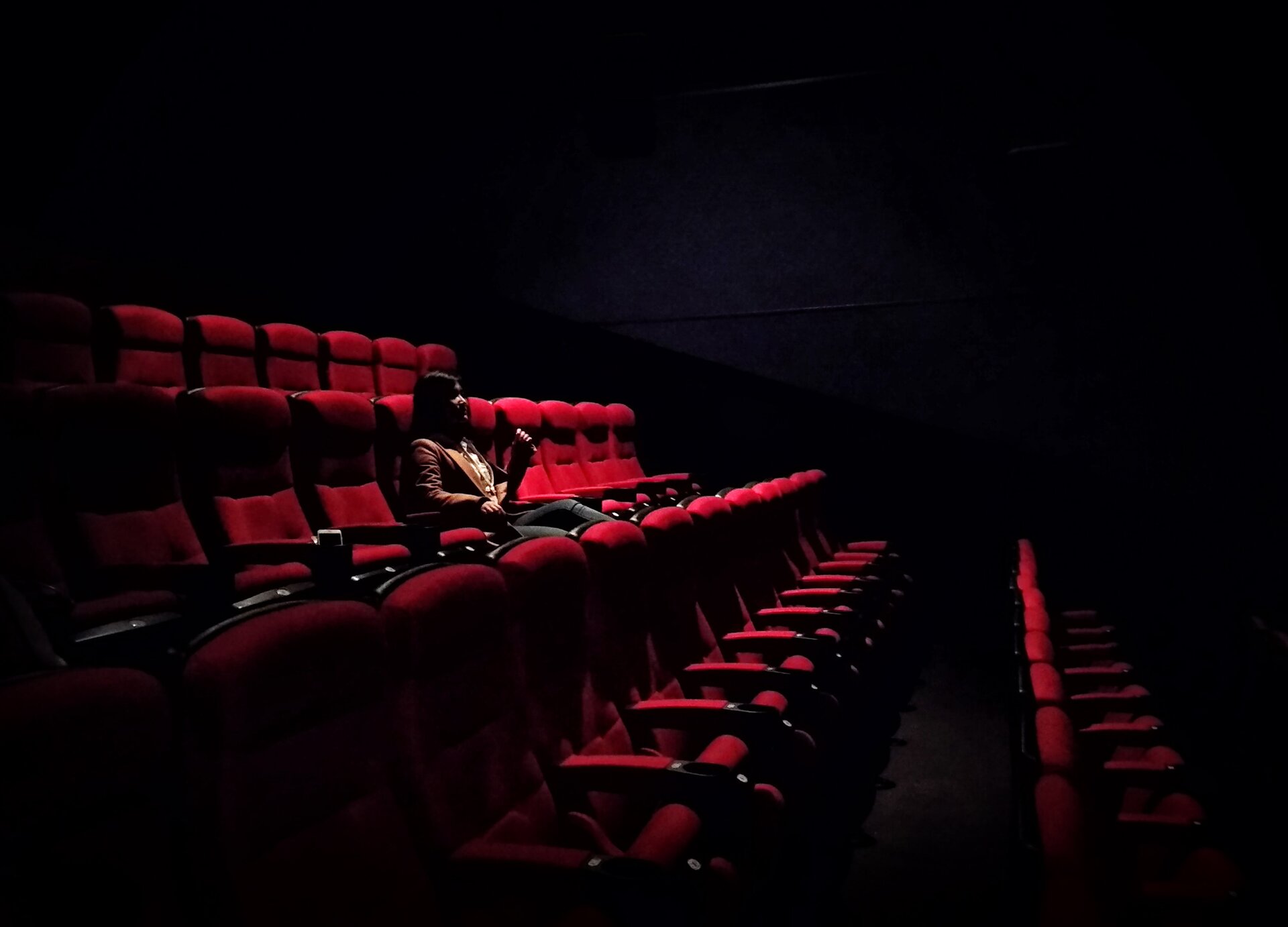 Zdjęcie przedstawia cztery rzędy czerwonych foteli na widowni w teatrze. W trzecim rzędzie od dołu widoczna jest siedząca kobieta, zwrócona w stronę sceny. 