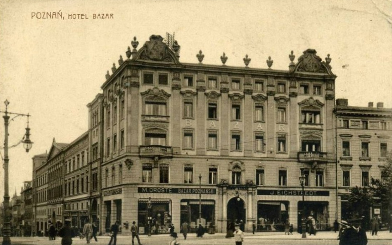 Ilustracja przedstawia budynek starego hotelu. Jest to budynek przypominający kubaturą dużą kamienicę o wysokości trzech pięter. Ma on bogato zdobioną fasadę. Na dole budynku znajdują się sklepy. Przed budynkiem znajduje się ruchliwa ulica po której spacerują ludzie. 