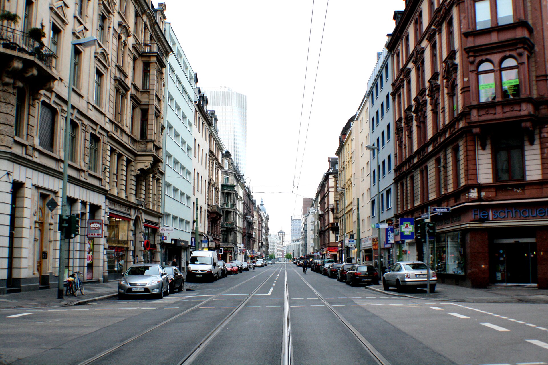 Ilustracja przedstawia ulicę miasta. Jest to prosta jezdnia z widocznymi dwoma torami tramwajowymi. Przy drodze stoją auta. Po obu stronach są wielopiętrowe kamienice o różnych kolorach elewacji. Nad jezdnią rozciąga się trakcja elektryczna dla tramwajów. W tle widać wieżowiec i jasne niebo.