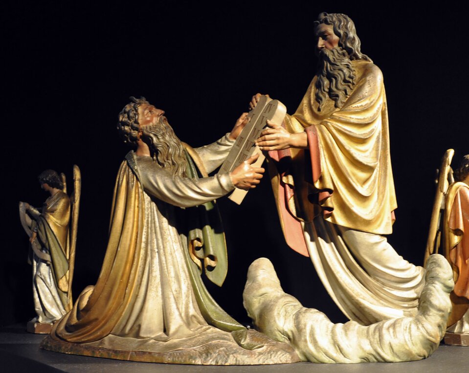 Ilustracja przedstawia posągi dwóch mężczyzn w obszernych szatach. Jeden klęczy, a drugi stojąc przekazuje mu dwie kamienne tablice, które mają kształt prostokątów zakończonych u górze półkolem. Obaj są starzy, mają długie włosy i brody. Po bokach stoją, odwrócone tyłem do sceny, dwa anioły.