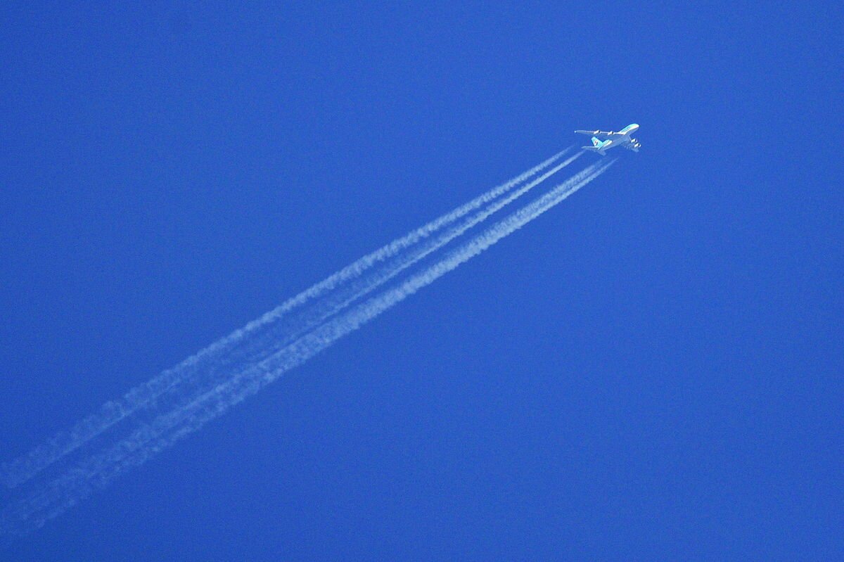 Zdjęcie przedstawia lecący samolot. Tłem jest niebieskie niebo. Samolot piały. Za samolotem widoczne białe linie przypominające chmury. Linie zaznaczają tor ruchu, po którym poruszał się samolot.