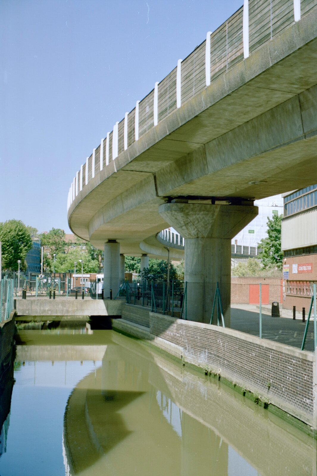 Na zdjęciu przedstawiony jest most zbudowany w centrum miasta. W tle widać budynki, a obok mostu przepływa rzeka. Most jest oparty na gęsto umieszczonych, betonowych belkach, z których jedna jest szersza od pozostałych.