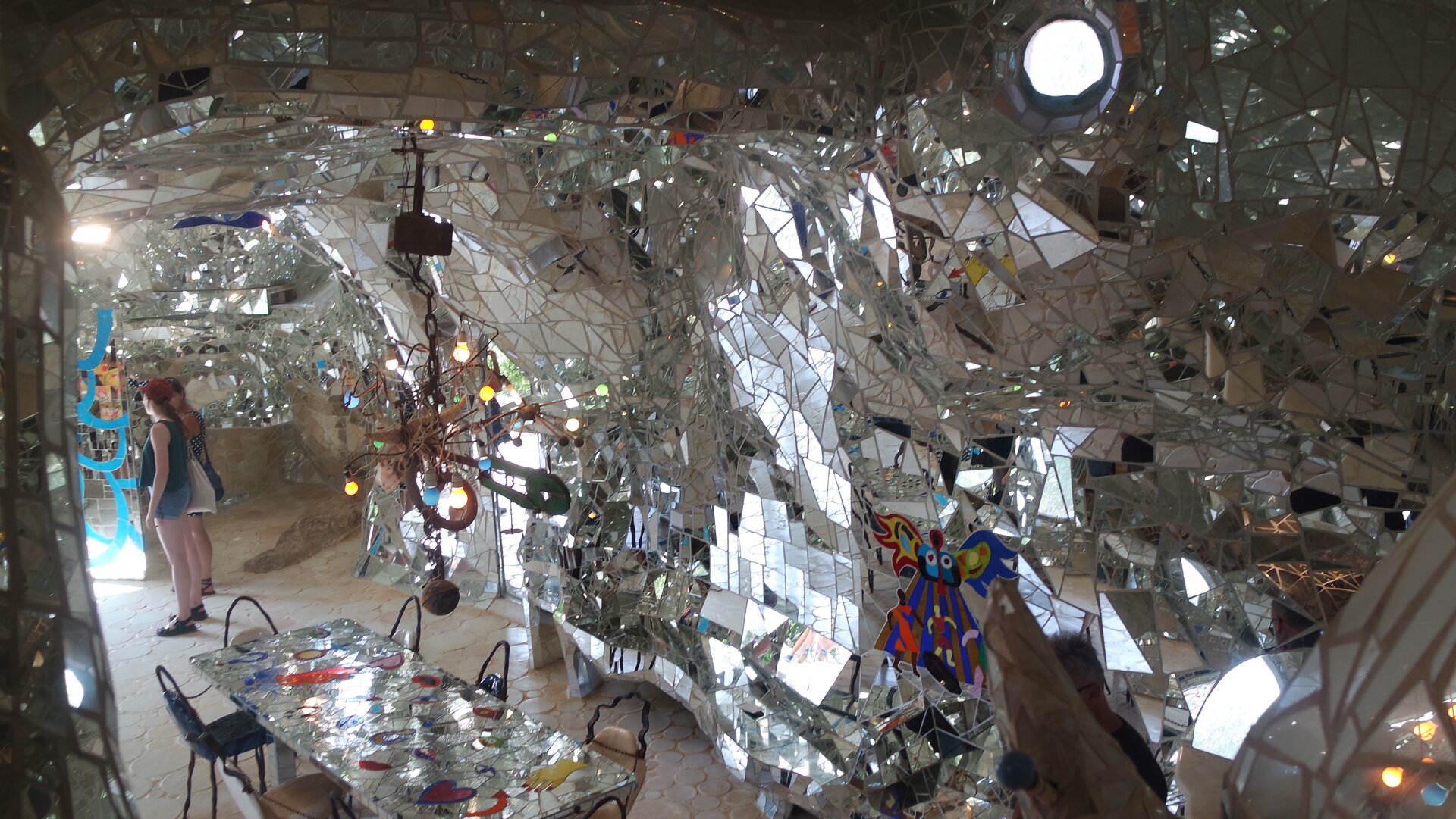 Ilustracja przedstawia pracę Niki de Saint Phalle. Jest to budynek-rzeźba Cesarzowa. Fotografia została wykonana w 2017 roku. Budowlę możemy udostępnić w Capalbio w Toskanii we Włoszech. Na zdjęciu widzimy wnętrze budowli. Ściany pokryte są mnóstwem małych elementów mozaiki. W budowli możemy dostrzec stół oraz krzesła, a także wiszącą lampę. Z lewej strony znajdują się dwie kobiety oglądają wnętrze. Autorką pracy jest francuska rzeźbiarka oraz malarka.