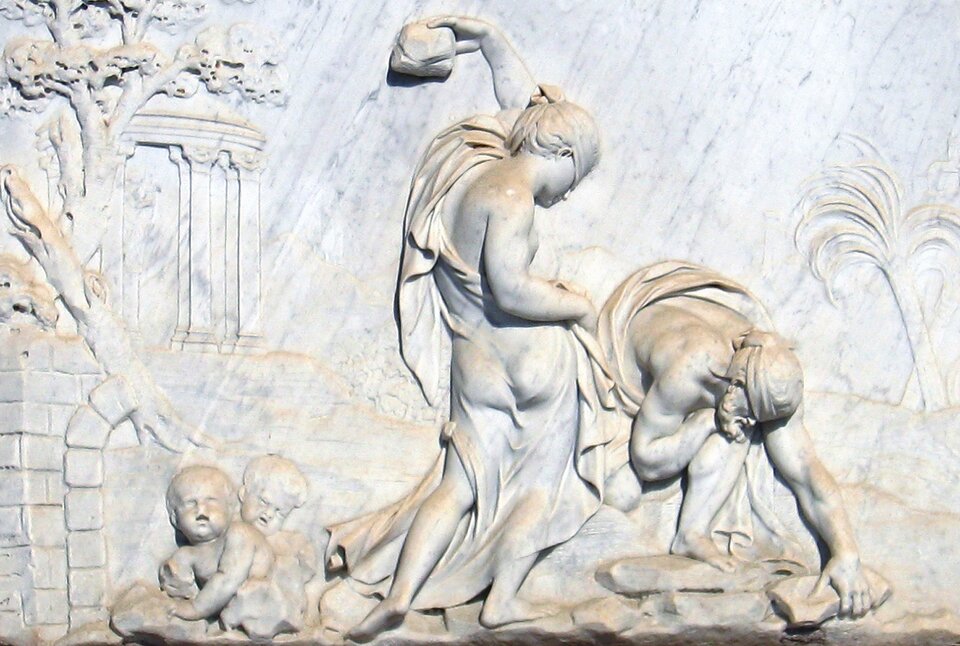 Fotografia przedstawia płaskorzeźbę wykonaną z jasnego kamienia. W centrum znajdują się dwie postaci. Kobieta ubrana w długą zwiewną szatę ukazana jest tyłem. Lewą rękę z kamieniem przechyla za siebie. Po jej prawej stronie znajduje się pochylony do ziemi mężczyzna. Jedną ręką podnosi z ziemi kamień. Za plecami kobiety na ziemi siedzi dwoje niemowląt. W lewym rogu płaskorzeźby znajduje się drzewo i fragment budowli.