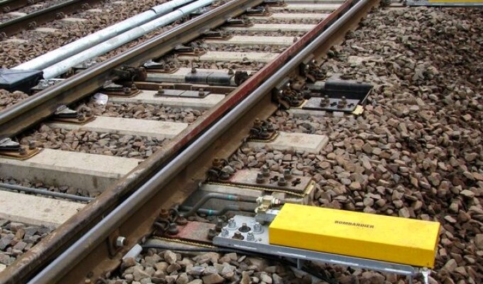 Zdjęcie przedstawia napęd zwrotnicowy. Jest to żółta skrzynka umiejscowiona obok torów w miejscu, gdzie jest możliwość przekierowania jadącego taboru na jeden z dwóch torów. Torowisko w tym miejscu składa się z czterech szyn, zewnętrznych — nieruchomych i wewnętrznych ruchomych. Zwrotnica jest połączona z ruchomymi częściami toru, dzięki czemu może zmieniać ich położenie. 