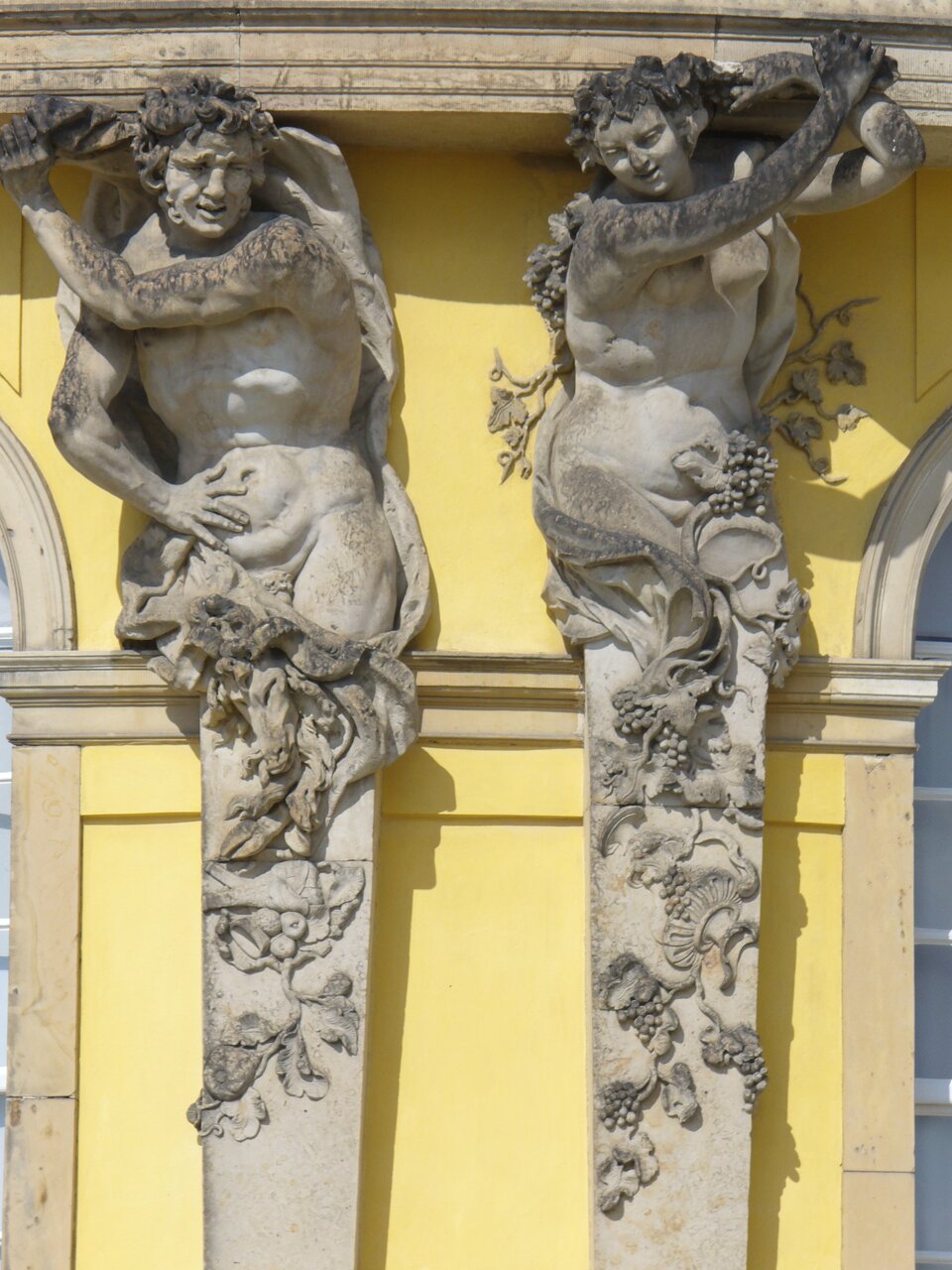 Ilustracja przedstawia zbliżenie na hermy elewacji południowej Pałacu Sanssouciw Poczdamie. Hermy zwieńczone są nagimi postaciami z orszaku Bachusa, owiniętymi zwojami winorośli.