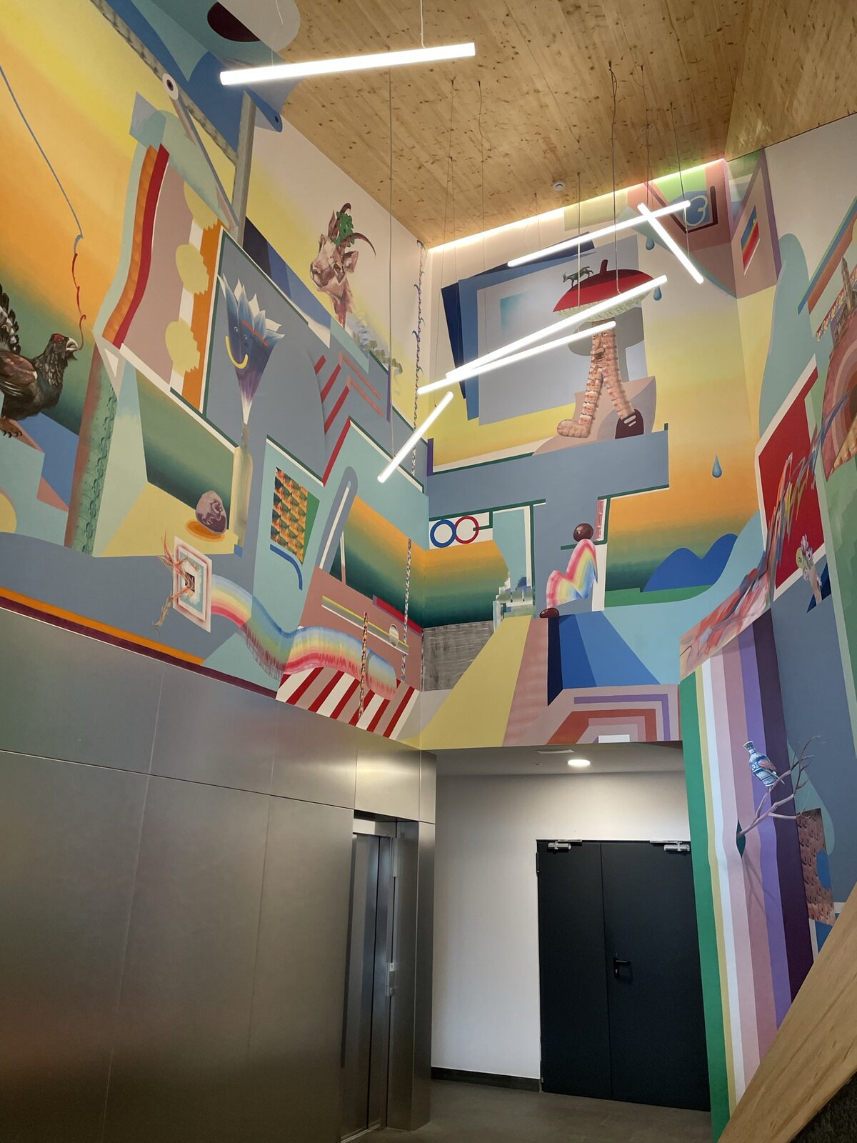 Zdjęcie przedstawia dekorację malarską na wysokiej ścianie szkolnego korytarza. Jest tu dużo motywów przedstawiających i abstrakcyjnych w płaszczyznach, które mają różne kształty. Kompozycja jest bardzo kolorowa, o barwach ciepłych, zimnych i gorących. Szeroki pas ściany od podłogi powyżej dwojga drzwi jest pomalowany jednolitym, jasnym kolorem. Kompozycja malarska obejmuje wysokie partie ściany ponad drzwiami i sięga aż do drewnianego sufitu. Z góry zwisa sześć lamp o kształcie prostych, długich rurek, a nad drzwiami świeci pojedyncza, okrągła lampa.