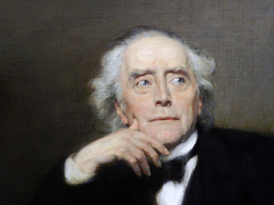 Obraz przedstawia twarz starszego mężczyzny. Jest ona owalna, z zaznaczonymi zmarszczkami. Mężczyzna ma krzaczaste brwi, wysokie czoło, długi nos. Jego włosy są siwe, falujące. Mężczyzna podpiera prawą dłonią prawy policzek. Jest ubrany w garnitur, koszulę i muchę.