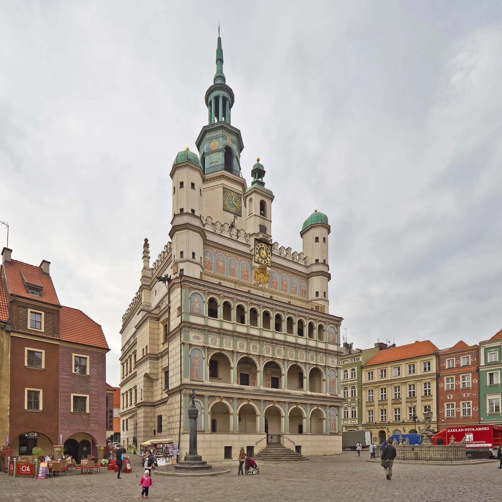 Zdjęcie przedstawia ratusz na rynku w Poznaniu. Ratusz to wysoki budynek, jego fasada z boku oraz w dolnej części jest jasnobeżowa i pomalowana ciemniejszym kolorem w kształt przypominający duże cegły. W bocznej części umieszczono także liczne, prostokątne okna. W przedniej części znajdują się schody do wejścia. Na prawie całej przedniej ścianie budynku umieszczono otwory w kształcie łuku, a między nimi niewielkie kolumny pomalowane na jasnozielony kolor. Przestrzenie nad łukami są pomalowane na jasne kolory, znajdują się na nich anioły, a skrajnie z obu stron przedniej ściany namalowano postacie rycerzy czy królów. Nad częścią z łukami stoi złota balustrada. Za nią namalowano więcej postaci, a między nimi znajduje się złoty zegar na czarnym tle. Na rogach budynku stoją kolumny. Kolumna na środku budynku, nad zegarem, mieści w sobie dzwon. Najwyższa część ratusza to ośmiokątna wieża, stojąca za kolumną z dzwonem. Ma ona na sobie kolejny zegar i jest zakończona strzelistą kopułą. Wokół budynku znajduje się kamienny deptak, po którym chodzą ludzie. Inne elementy to mała kolumna z rzeźbą, restauracje, kamienna fontanna oraz budynki mieszkalne.