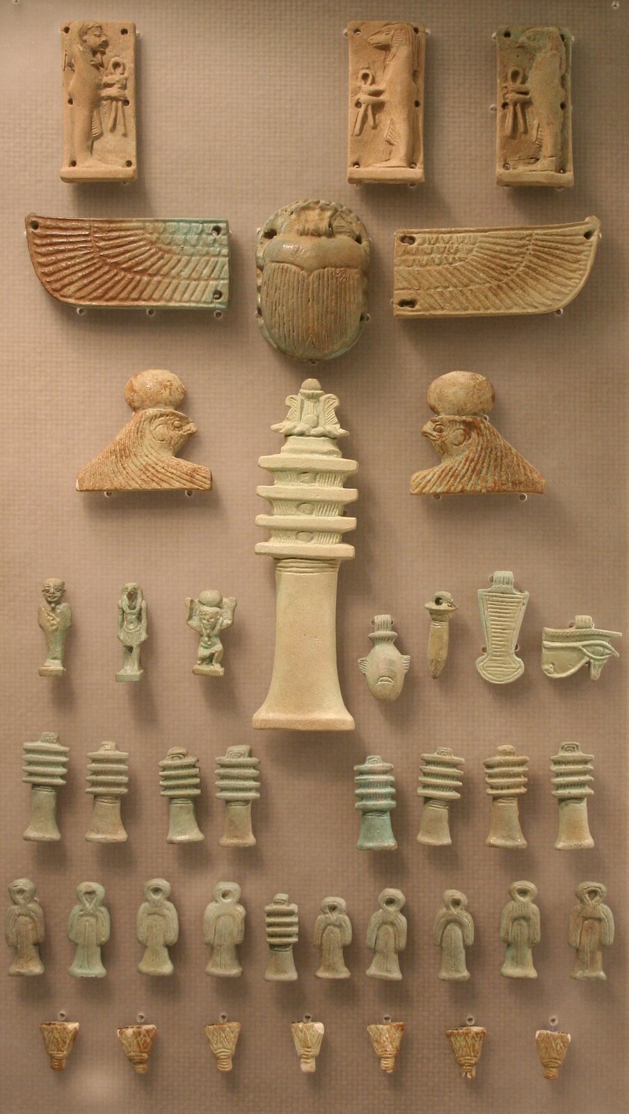 Zdjęcie przedstawiające zestaw egipskich amuletów. Od góry amulety z wizerunkami faraona, bogów, skarabeusza, skrzydeł, boga Ra, wizerunek z głową sokoła a także szereg innych amuletów.