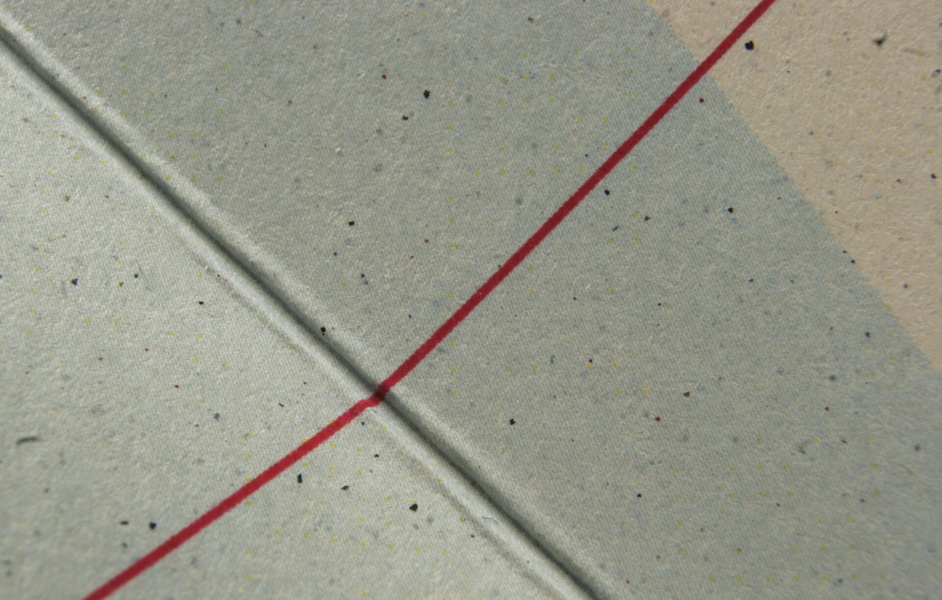 Zdjęcie przedstawia bigowanie okładki. Widoczny jest szary grzbiet okładki, pod kątem, z wyraźnie zaznaczonym zagłębieniem, czyli bigowaniem. Poprzecznie do niego biegnie cienka czerwona linia.