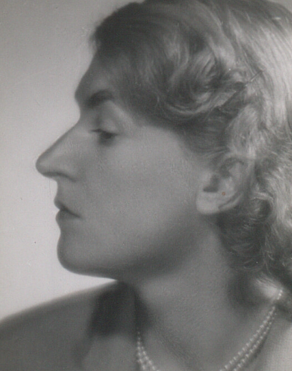 Zdjęcie przedstawia młodą kobietę. Ukazany jest jej lewy profil. Kobieta ma długie, jasne, falujące włosy. Na szyi naszyjnik z pereł.