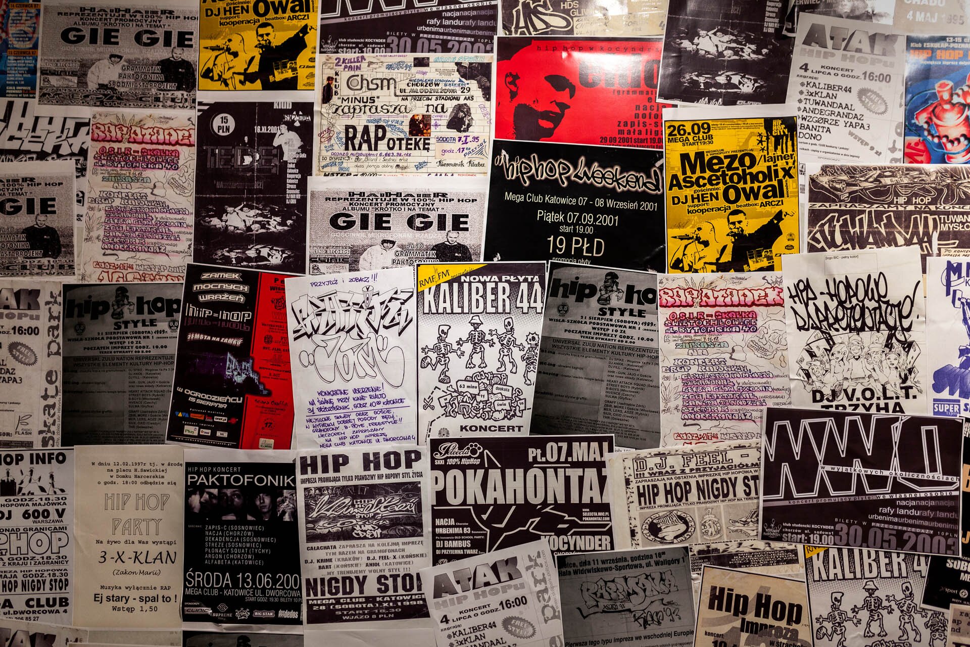 Fotografia przedstawia widok na fragment ściany pokrytej plakatami promującymi koncerty wykonawców hip‑hopowych, między innymi: Kaliber 44, Pokahontaz, Mezo, WWO, Nagły atak spawacza, Paktofonika. Większość jest czarno‑biała. Na ścianie wyróżniają się dwa żółte plakaty oraz dwa czerwono‑czarne. Plakaty umieszczone są bardzo blisko siebie – ich krawędzie na siebie nachodzą. Są prostokątne, niektóre pionowe, niektóre poziome.